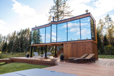 Çağdaş ahşap bir ev, geniş yansıtıcı pencereler, geniş bir güverte, açık hava koltukları, ve orman ortamında uzun ağaçlar..