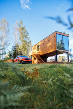 Ağaçları yansıtan büyük camlı camlı camları olan minimalist ahşap bir ev yanına park edilmiş kırmızı bir araba ve güvertede açık hava oturma alanı..