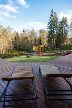 Bir güvertedeki iki ahşap şezlong pencereli modern bir ahşap yapıya bakıyor, yakınlarda bir şömine ve ağaçlarla birlikte çimlerin üzerine kurulmuş..