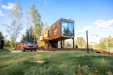 Cam cepheli ahşap bir kulübe ayaklıkların üzerinde duruyor, açık hava oturma alanı var. Yakınlarda huş ağaçları ve çimlerle çevrili turuncu bir araba var..
