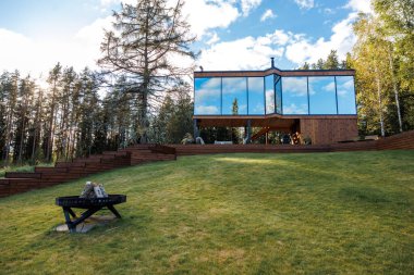 Yansıtıcı camlı camlı camları olan modern ahşap bir ev, uzun ağaçlı çimenli bir yamaca kurulmuş, şömine çukuru ve yokuş yukarı giden tahta bir merdiven..