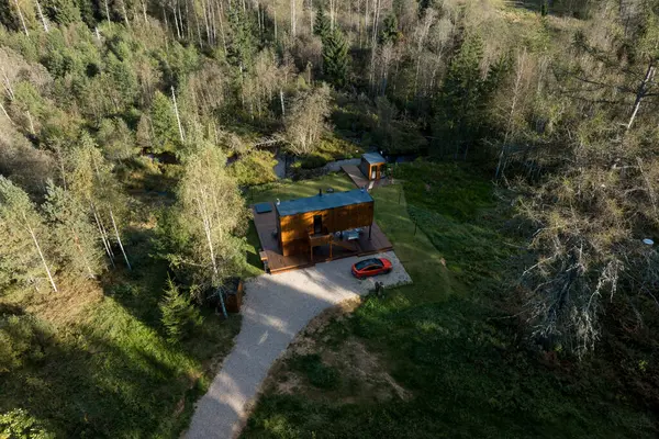 Havadan bakıldığında, yüksek ağaçlarla çevrili, düz çatılı, modern bir ahşap kulübe görülüyor. Yapının yakınında bir çakıl yolu ve kırmızı bir araba görünüyor..