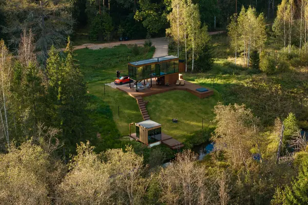Daha küçük bir yapı, kırmızı bir araç, toprak bir yol ve doğal bir akarsu ile kaplı, ahşap bir platformda modern cam duvarlı bir ev..