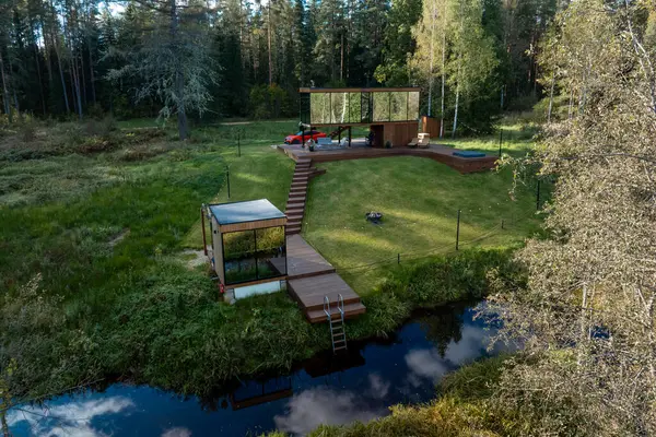 Camdan duvarları, ahşap bir güvertesi, açık hava koltukları, jakuzisi ve çim ve uzun ağaçlarla çevrili bir deresi olan iki katlı modern bir inziva yeri..