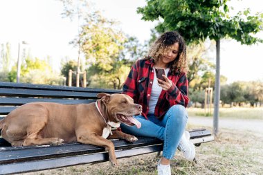 Parkta oturmuş safkan köpeğini okşayan kıvırcık saçlı genç bir kadın. Telefonuyla fotoğraf çekiyor..