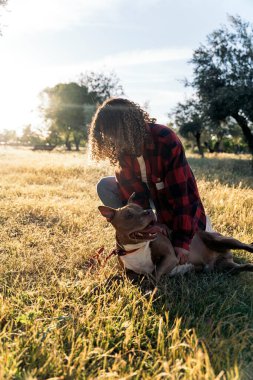 Kıvırcık saçlı genç kadın parkta köpeğini okşayarak eğleniyor..