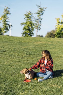 Afrikalı bir kadın köpeğiyle parkta oturuyor ve cep telefonunu kullanıyor..