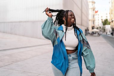 Havalı örgüleri olan, gülümseyen ve sokakta yürüyen neşeli Afrikalı genç bir kadın..