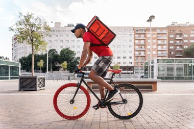 African delivery man wearing cap riding his bike in the city in his way to deliver a package. clipart