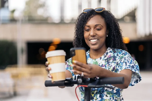 Happy african woman with curly hair using her mobile phone and holding coffee cup. She has an electric scooter.