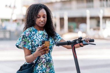 Elektrikli scooterlı mutlu siyah kadın sokakta yürüyor ve cep telefonunu kullanıyor..