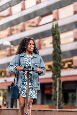 Genç siyah kız, güneşli bir günde şehirde elektrikli scooterıyla geziyor..