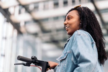 Güzel, kıvırcık saçlı Afrikalı bir kadın şehirde elektrikli scooterıyla eğleniyor..