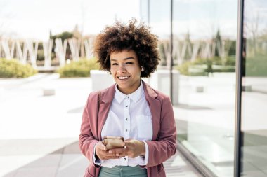 Açık havada cep telefonunu kullanan afro saçlı güzel siyahi bir iş kadını. Resmi kıyafetler giyiyor..