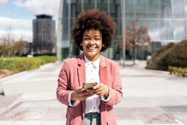 Güzel siyah iş kadını, afro saçlı cep telefonunu kullanıp kameraya bakıyor. O, şehrin bir iş alanında yürüyor..