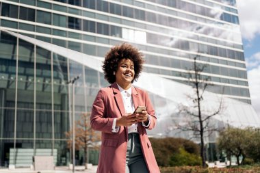 Afro saçlı, gülümseyen ve dışarıda cep telefonunu kullanan mutlu bir iş kadını..
