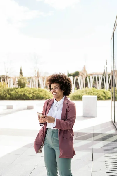 Pretty black business woman with afro hair using her mobile phone outdoors. She is wearing formal clothes.
