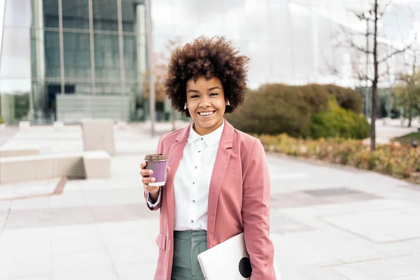 Pretty black business woman with afro hair holding her laptop and a coffee cup looking at camera. She is walking in a business area of the city.