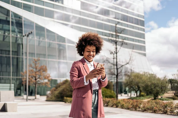 Happy black business woman with afro hair smiling and using her mobile phone outdoors in a business area.