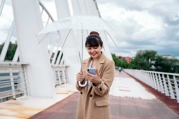 Szczęśliwa Młoda Kobieta Trzyma Parasol Pomocą Telefonu Komórkowego Ulicy Podczas — Zdjęcie stockowe