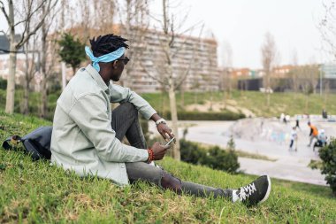 Havalı Afrikalı adam çimlerin üzerinde oturuyor ve cep telefonunu kullanıyor..