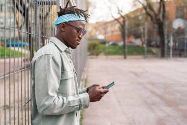 Genç ve havalı siyahi bir adam çitte duruyor ve cep telefonunu kullanıyor..