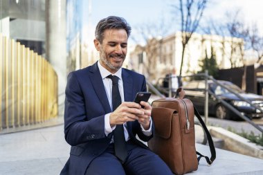 Tam vücut olgun takım elbiseli patron deri çantanın yanında oturuyor ve güneşli bir günde cep telefonunu karıştırıyor.