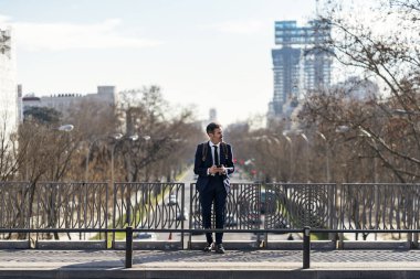 Şık takım elbiseli erkek girişimci güneşli bir sabahta caddede duruyor ve cep telefonuna bakıyor.