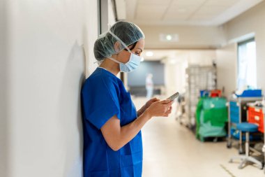 Doctor leaning against the wall using the mobile phone in a hospital ward alone