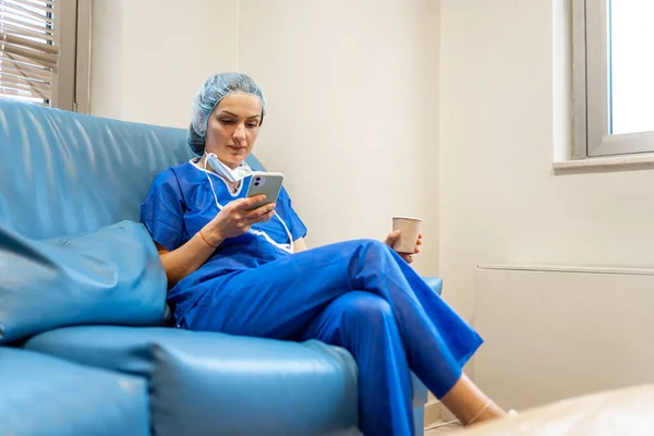 Nurse using the mobile phone during her break sitting on a sofa with a coffee