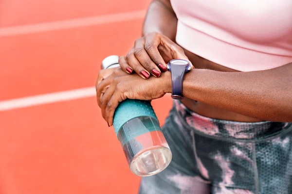 陸上競技場に立つアフリカ系アメリカ人のスプリンターのストック写真水のボトルで彼女の時計を見て — ストック写真