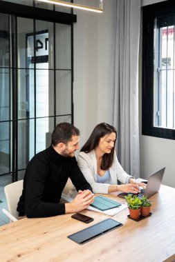 Genç adam ve kadın modern ofiste masada oturuyor ve proje üzerinde çalışıyorlar.