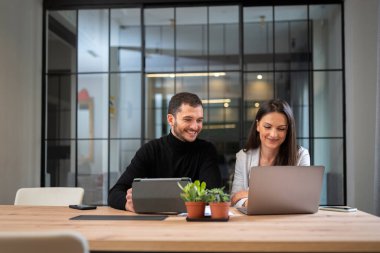 Genç adam ve kadın modern ofiste masada oturuyor ve proje üzerinde çalışıyorlar.