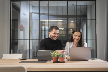 Genç adam ve kadın modern ofiste masada oturuyor ve proje üzerinde çalışıyorlar.