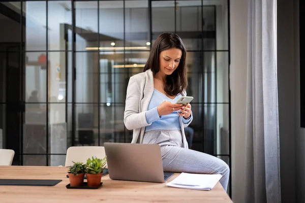 stock image Concentrated young woman in casual outfit sitting at table with laptop and messaging on mobile phone while working on project in the office