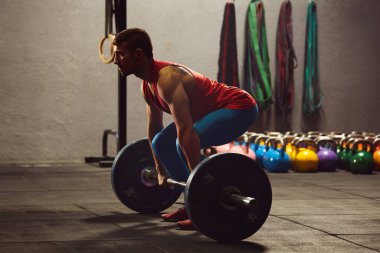 Spor salonunda çömelmiş halter kaldıran yetişkin bir adamın stok fotoğrafı. Spor giyim kuşamı giyiyor. Arka planda halterler var..