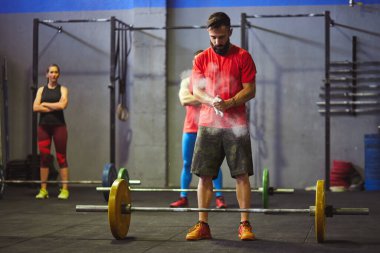 Stock photo of an adult man in a gym chalking his hands. There is a barbell in front of him and people behind him. They are wearing sportswear. clipart