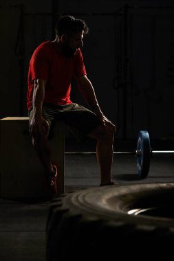 Spor salonunda oturan ve dinlenen yetişkin bir adamın stok fotoğrafı. Önünde bir traktör lastiği ve arkasında bir halter var. Spor giysisi giyiyor..