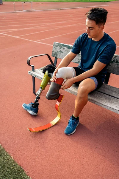 Stock photo of disabled man athlete sitting in a bench and putting on his leg prosthesis.