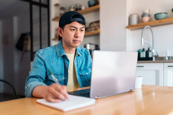 Asiatischer Mann Schreibt Notizbuch Während Laptop Der Küche Benutzt — Stockfoto