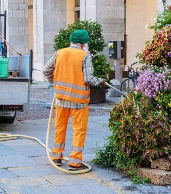 Cuneo, İtalya - 14 Temmuz 2022: Yeşil sarıklı ve turuncu işçi elbiseli bir adam İtalyan şehrindeki saksılarda çiçekleri suluyor