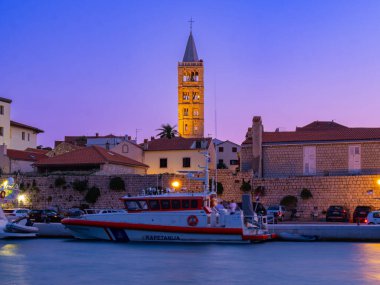 Rab, Hırvatistan - 19 Ağustos 2023: Alacakaranlıkta Kapetancija adlı bir devriye teknesiyle sakin bir sahil sahnesi, bu da tarihi binalarla çevrili küçük bir limanda sahil güvenlik anlamına geliyor ve uzun bir çan kulesi, değişen bir gökyüzü altında. Önde ve sakin.