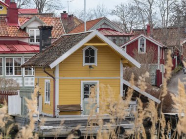 Vaxholm, Sweden - 11 April 2023: Charming yellow cottage by serene water and other houses in the background, surrounded by lush greenery, exuding tranquility and beauty. clipart