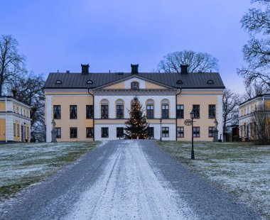 Nykvarn, İsveç - 23 Ocak 2023: Önünde karlı bir manzarayla çevrili bir Noel ağacı olan büyük tarihi bir bina. Binanın birden fazla penceresi ve merkezi girişi olan simetrik bir tasarımı var. Gökyüzü bulutlu ve yer de... 