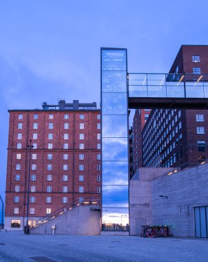 Stockholm, İsveç - 3 Mart 2023: İki yapıyı birbirine bağlayan cam bir köprüye sahip modern bir bina. Alt kısımda beton taban yer alırken, üst kısımda çok sayıda pencereli bir tuğla ön cephe yer alıyor. Sahne alacakaranlıkta geçer, yansıtır.