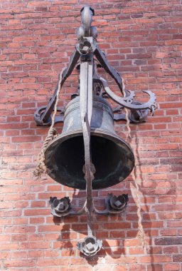 Süslü, yıpranmış bir çan tuğla bir duvara monte edilmiş, dekoratif metal işçiliği ve iplerle tamamlanmış.