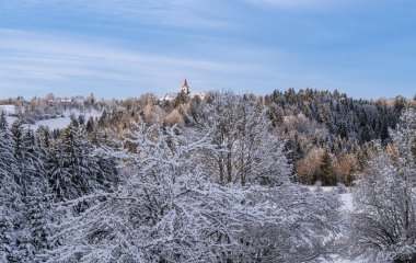 Karla kaplı ağaçların ve açık mavi gökyüzünün altındaki tepede uzak bir kilisenin bulunduğu manzaralı bir kış manzarası..