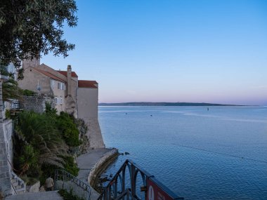 Rab, Hırvatistan - 10 Ağustos 2024: Gün doğumunda deniz kenarında tarihi bir taş binanın yer aldığı sakin bir kıyı manzarası. T
