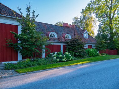 Nacka, İsveç - 6 Eylül 2025: Çatı kiremit kaplı, yemyeşil ve çiçek açan çalılarla çevrili büyüleyici kırmızı ahşap bir ev. Sahne sessiz bir caddede geçiyor, sabah ışığıyla aydınlatılıyor..