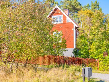 Vaxholm, İsveç - 6 Eylül 2024: yemyeşil ve ağaçlarla çevrili büyüleyici kırmızı ahşap bir ev. Evin önünde kırsal ahşap bir çit bulunan eğimli bir çatı ve beyaz bir süsleme yer alıyor. Ön planda bir posta kutusu görülebilir, serene eklenir